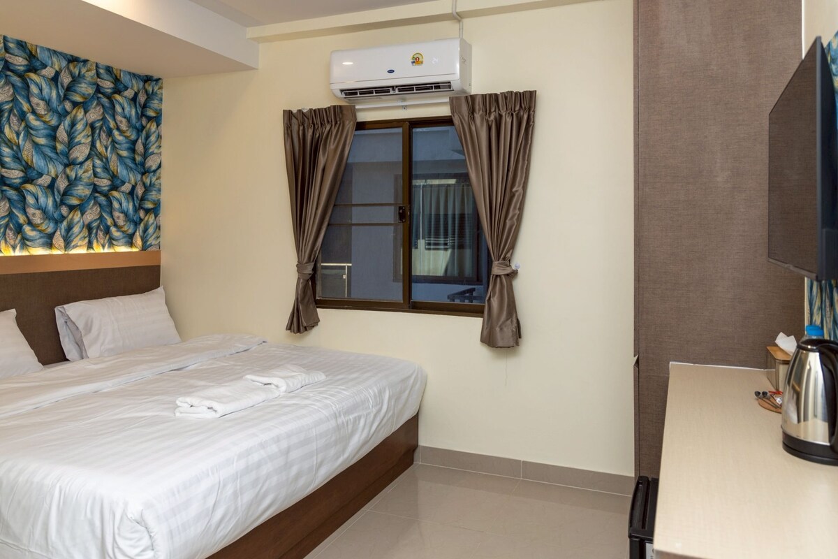 A hotel room with a bed, a window with curtains, an air conditioner, a flat-screen TV, and a kettle on the table.