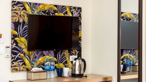 Une chambre d’hôtel dotée d’un grand téléviseur à écran plat fixé au mur, d’un bureau en bois, d’une bouilloire, de tasses et de bouteilles d’eau.