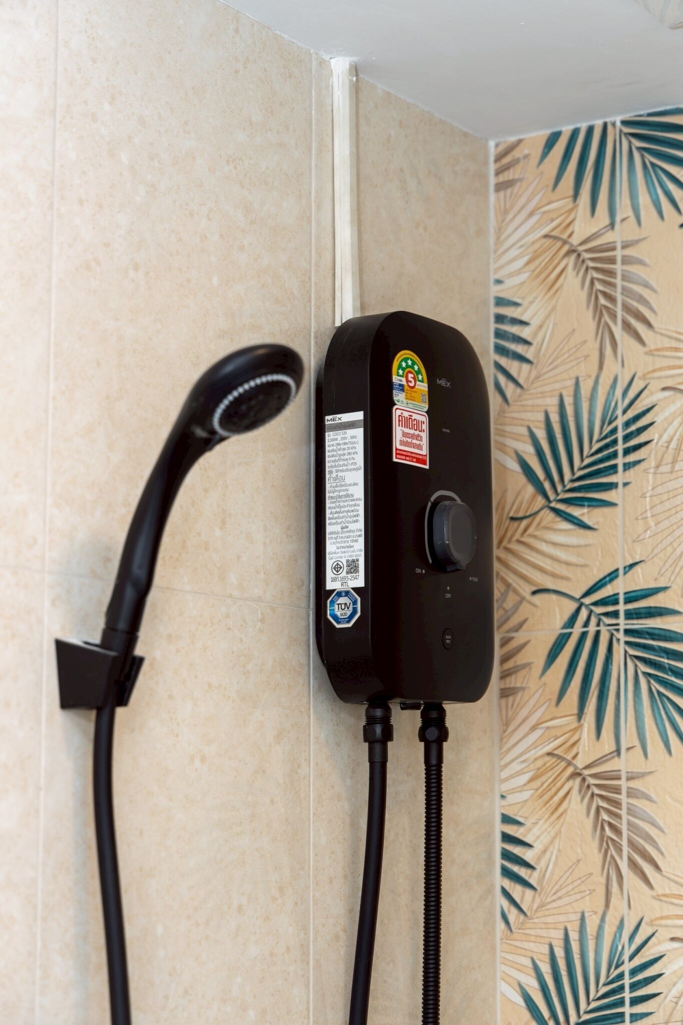 Un pommeau de douche et un panneau de commande fixés sur un mur recouvert de carreaux décorés d’un motif de feuilles tropicales.