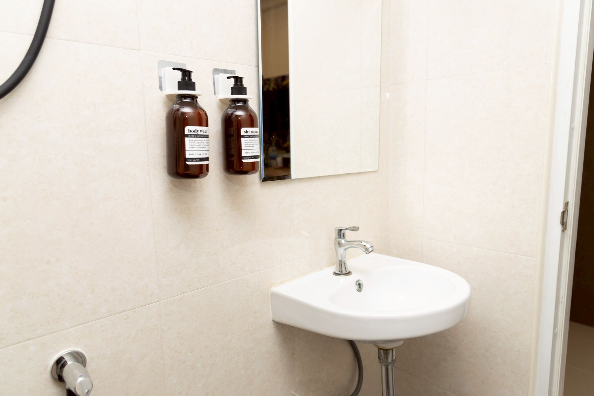 A bathroom with a white sink, two brown bottles labeled "body wash" and "shampoo" mounted on the wall, and a mirror above the sink.