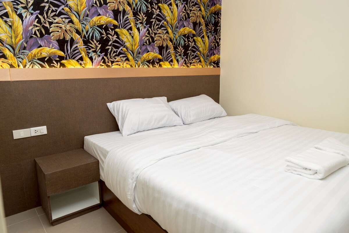 A neatly made bed with white linens, a small bedside table, and a wall with a tropical leaf pattern.