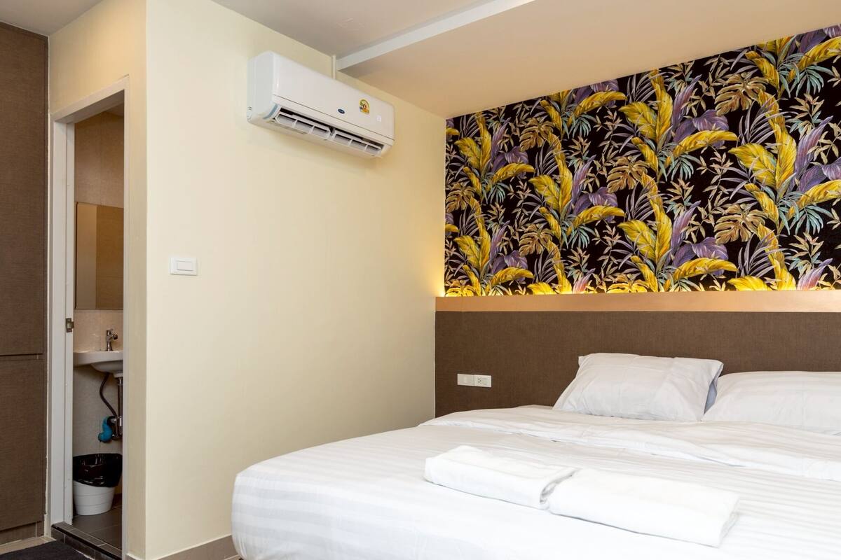 A hotel room with a bed, a wall-mounted air conditioner, a bathroom with a sink and toilet, and a decorative wall with a tropical leaf pattern.