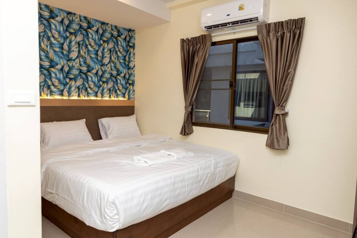 A neatly made bed with white linens, a wall-mounted air conditioner, and a window with curtains.