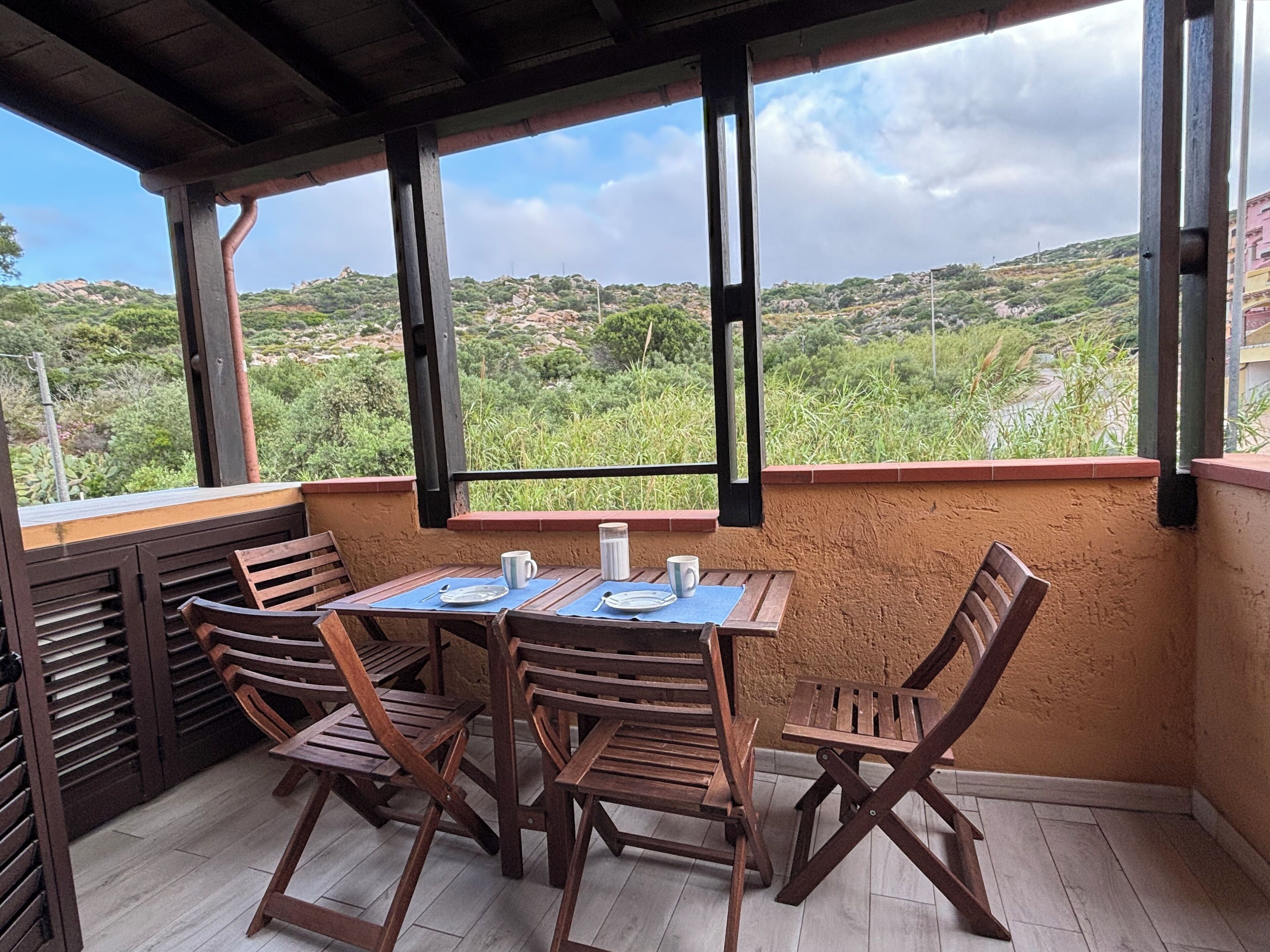 A covered outdoor seating area with a table and chairs, overlooking a hilly landscape.