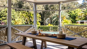 A wooden table with a glass dome centerpiece, a bottle, and a glass of wine on a balcony overlooking a pool and lush greenery.