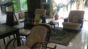 Ein Wohnzimmer mit Schaukelstuhl, Sofa und Couchtisch. Üppige Zimmerpflanzen und ein Fenster mit Jalousien.