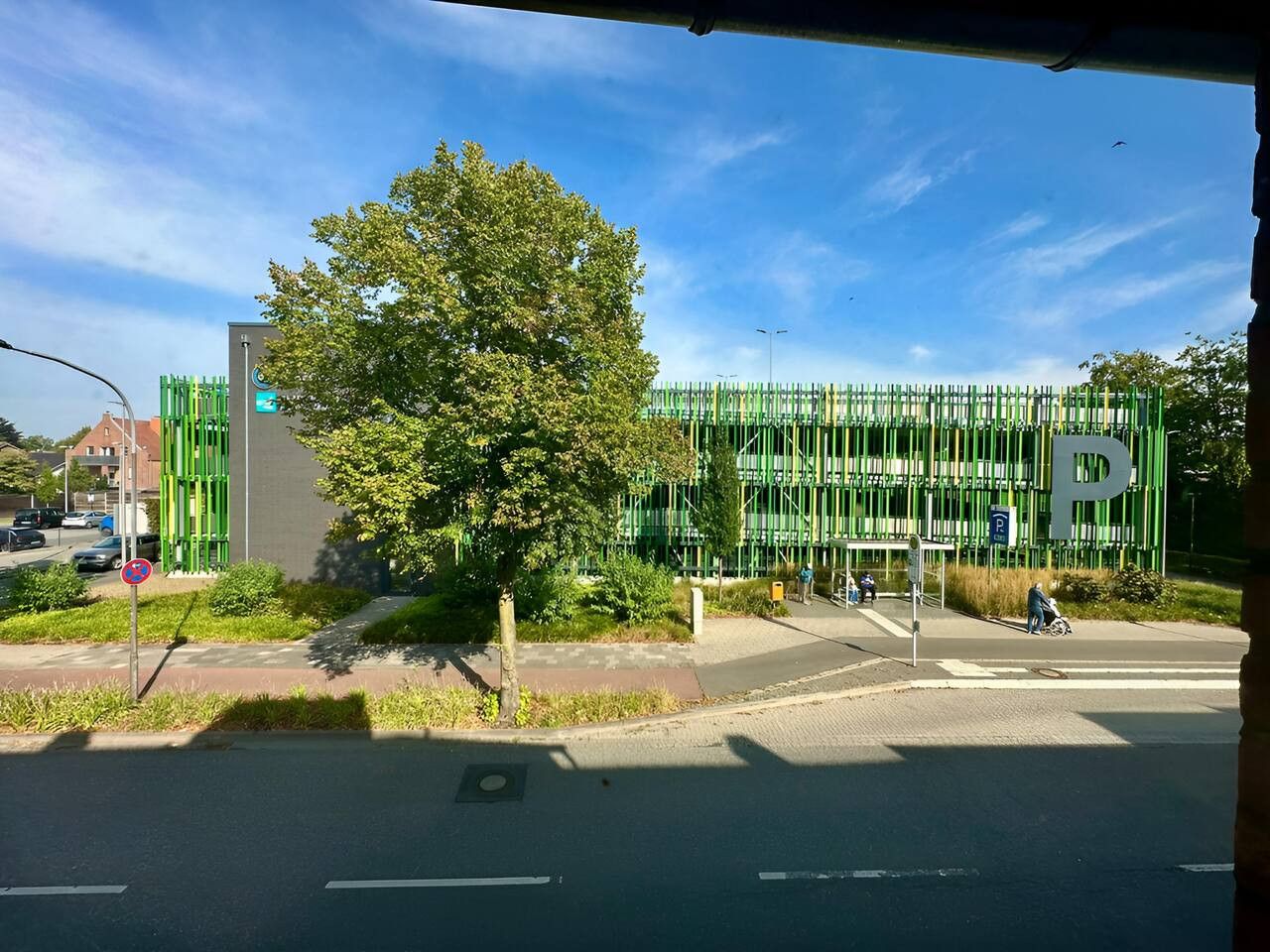 Straatbeeld met een groot groen gebouw met een 'P'-bord, wat een parkeerplaats aangeeft.