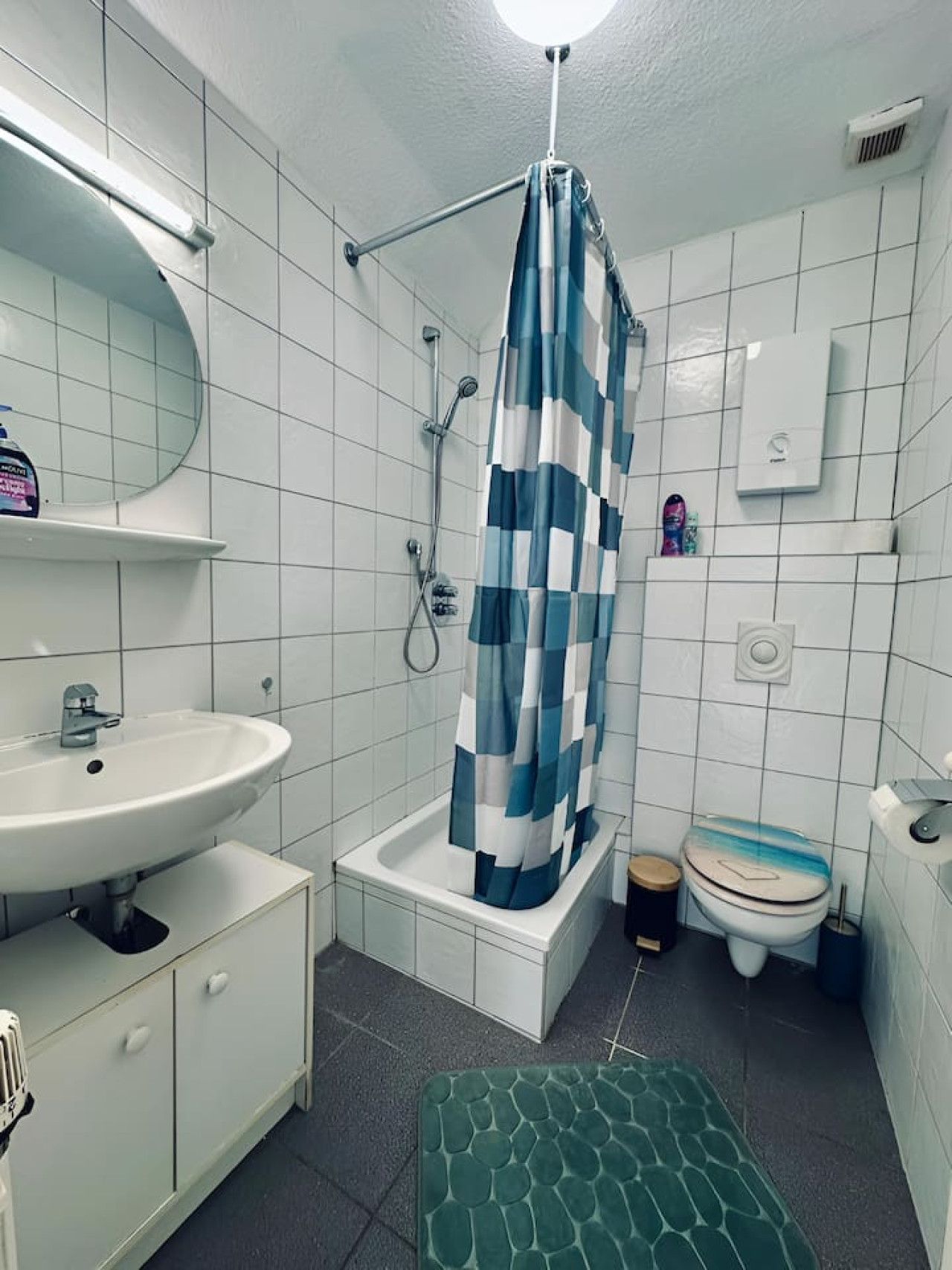 Ein Badezimmer mit einem weißen Waschbecken, einer Dusche mit blau-weißer Karomuster-Duschvorhang, einer Toilette und einem grün gemusterten Teppich auf dem Boden.