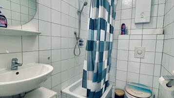 Een badkamer met een witte wastafel, een douche met een blauw-wit geruit gordijn, een toilet en een vloerkleed met een groen patroon.