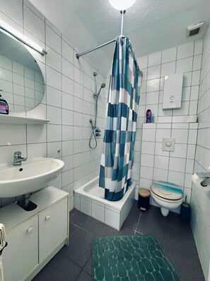 Een badkamer met een witte wastafel, een douche met een blauw-wit geruit gordijn, een toilet en een vloerkleed met een groen patroon.