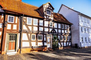 Ein traditionelles Fachwerkhäuschen mit einer hölzernen Bank vor der Tür, einer Tür mit einer Hausnummer und einem kleinen Baum.