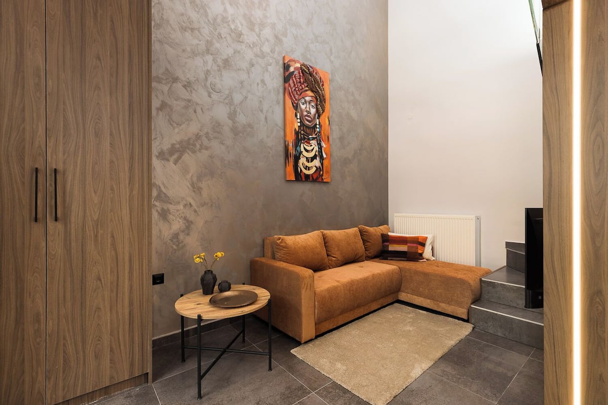 A modern living room with a wooden wardrobe, a brown sofa, a small round table with a vase and a bowl, and a painting on the wall.