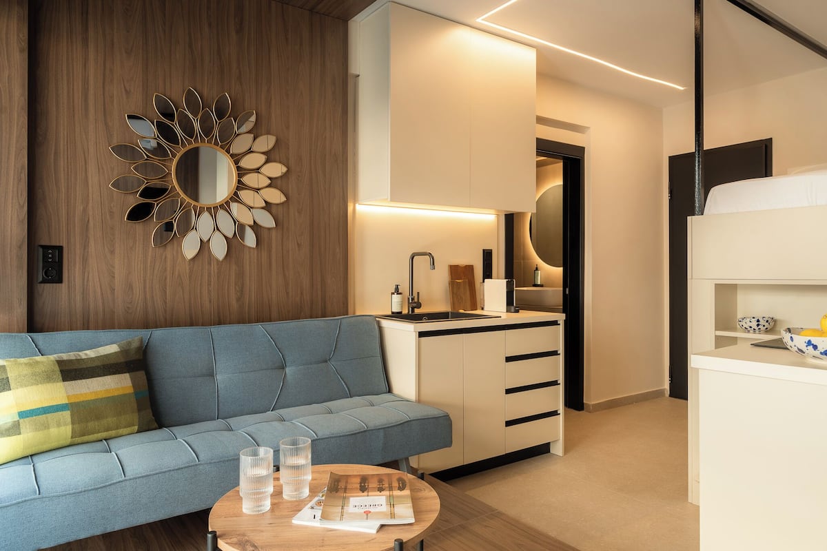 A modern hotel room with a blue sofa, a wooden accent wall, a kitchen area, and a bed.