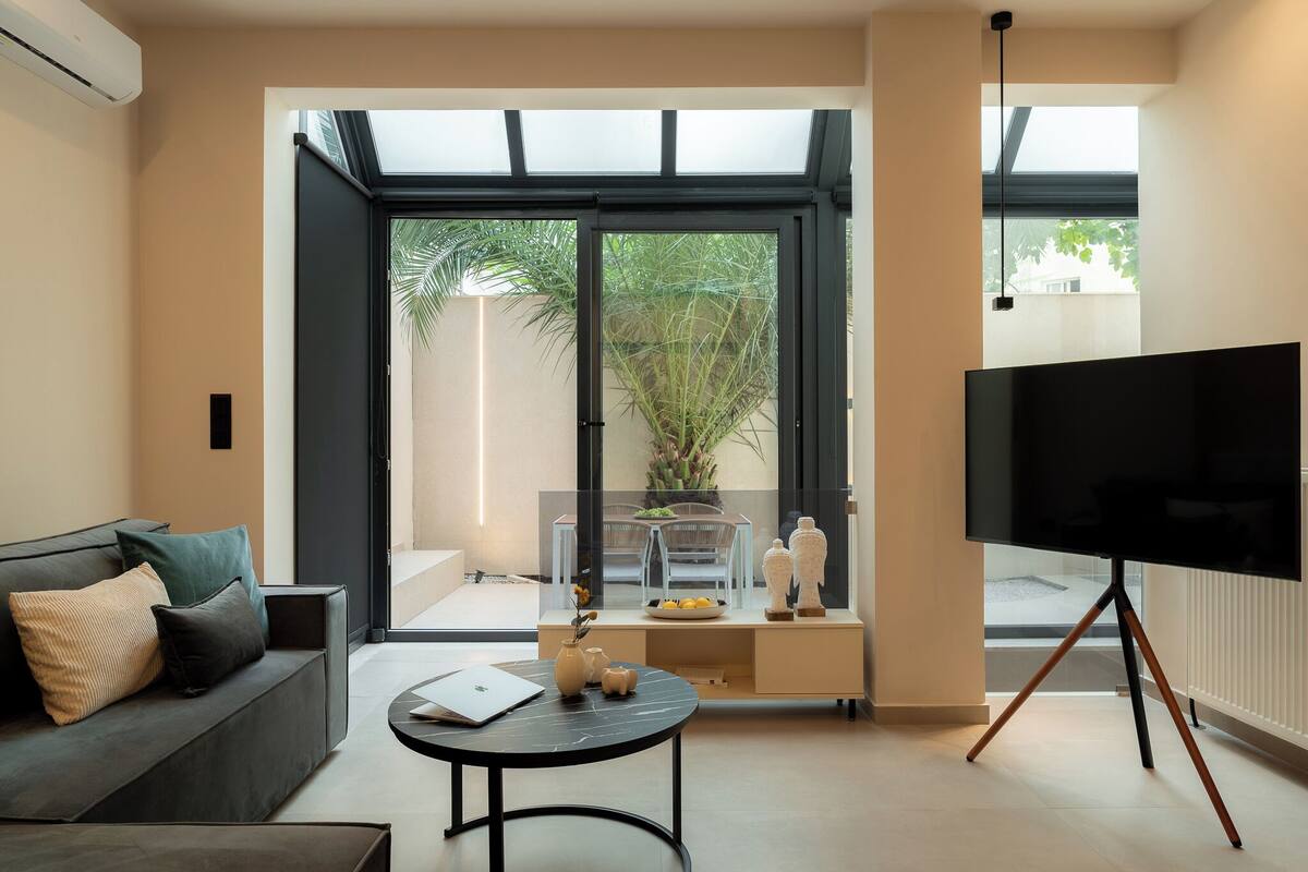 A modern living room with a sofa, a coffee table, a TV on a tripod stand, and a large window with a view of greenery outside.