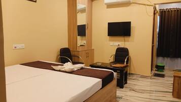A hotel room with a bed, two chairs, a desk, a flat-screen TV, an air conditioning unit, and a mirror.