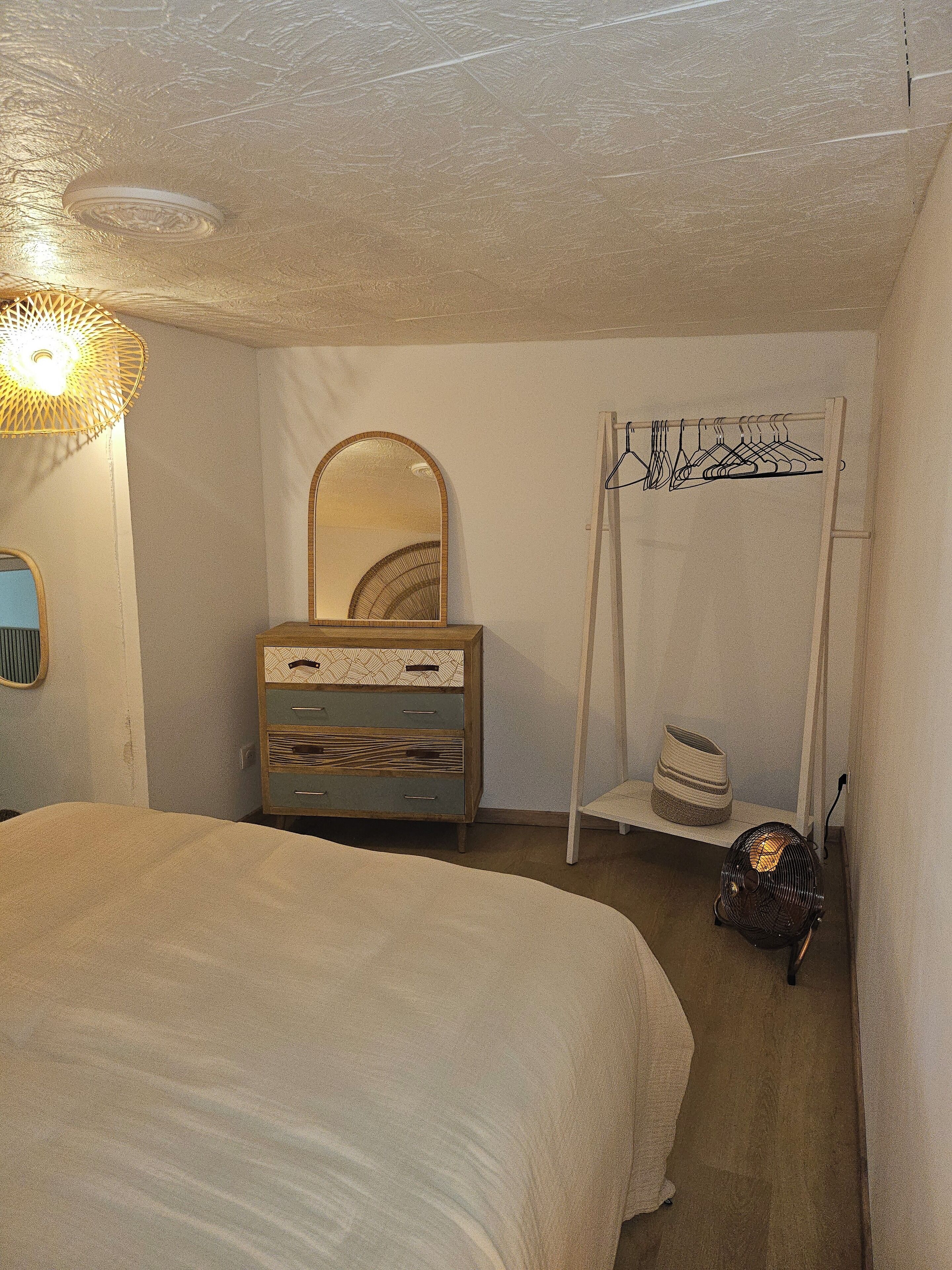 Een slaapkamer met een groot bed, een dressoir met lades, een spiegel, een kledingrek en een klein plankje met manden.