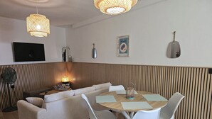 A modern living room with a sofa, dining table, and chairs. There is a television mounted on the wall, a lamp on the table, and a decorative item on the wall. - 54m² cocooning apartment in the center of St Cannat (Saint-Cannat)