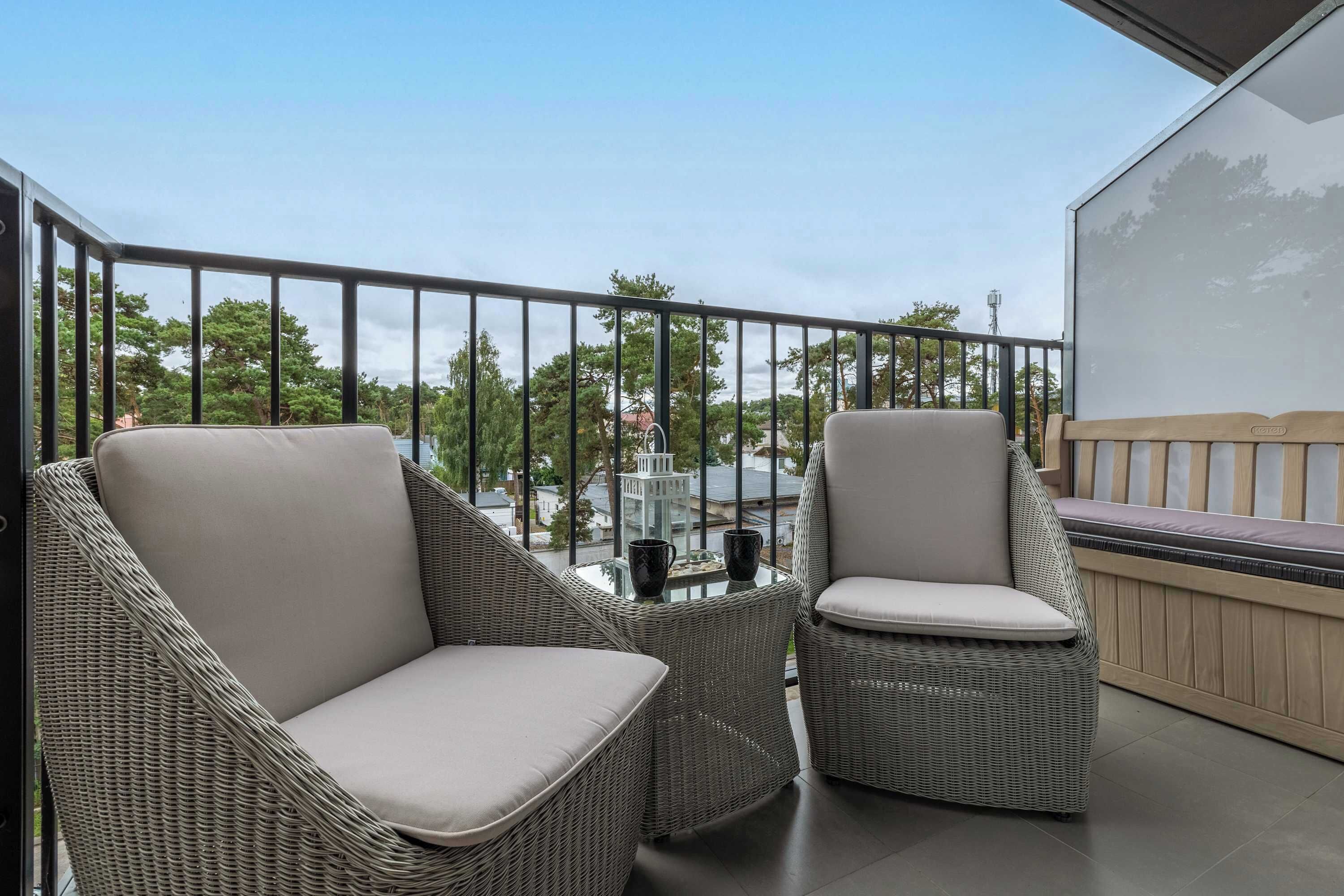 Un balcón con mobiliario de mimbre, una mesa de vidrio y vistas a árboles y edificios.