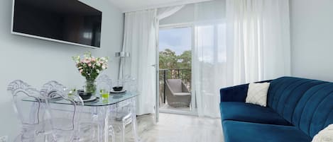 Een moderne woonkamer met een glazen eettafel, transparante plastic stoelen, een blauwe bank en een aan de muur bevestigde televisie.