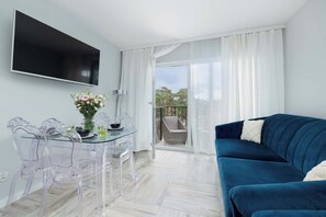Uma sala de estar moderna com uma mesa de jantar de vidro, cadeiras de plástico transparente, um sofá azul e uma TV fixada na parede.