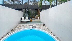 A modern outdoor pool area with a clear blue pool, surrounded by white pebbles and a white wall. There is a seating area with a sofa and cushions, and a wooden deck above the seating. - Luxury Private Pool Loft Villa (Canggu)