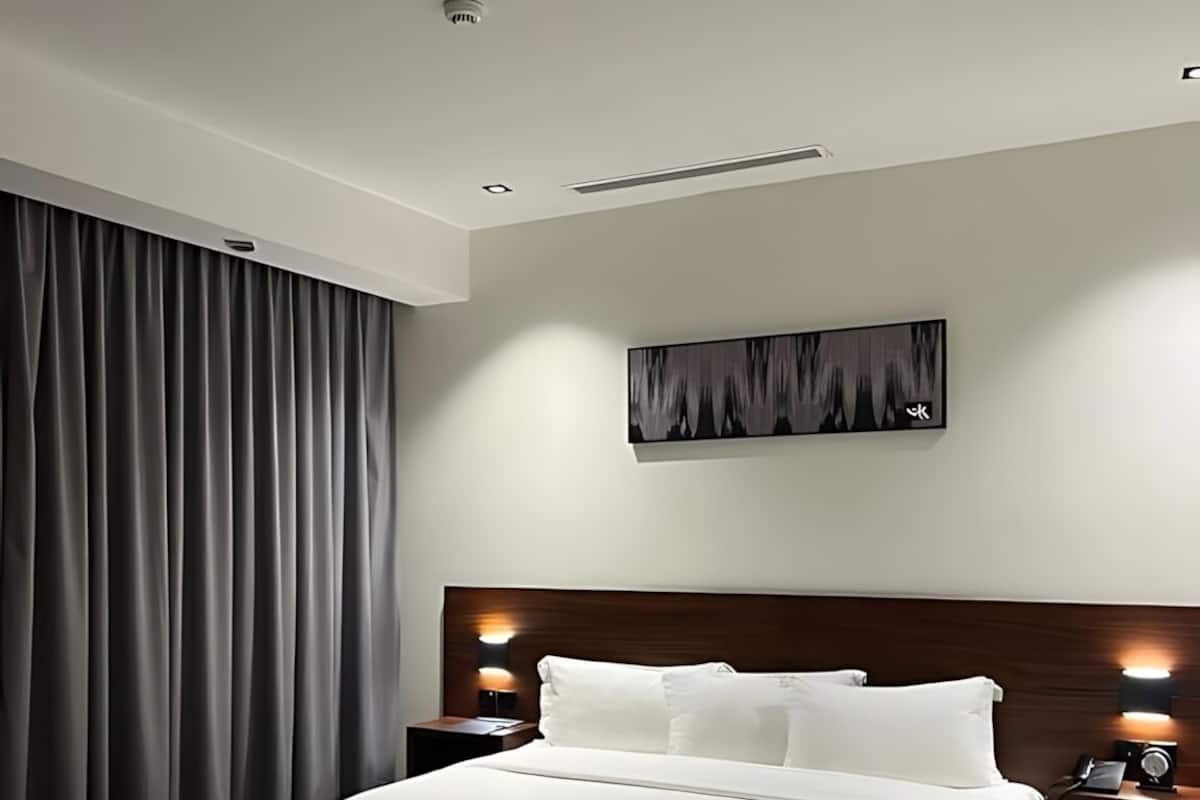 A modern hotel room with a large bed, a headboard with integrated lighting, a bedside table with a lamp, a gray ottoman, and a painting on the wall.