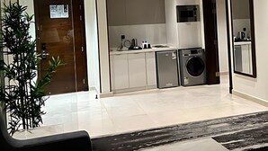 Una sala moderna con sofá, mesa de centro y un ventanal grande. Se observa una zona de cocina con fregadero, estufa y microondas. Una planta se ve en la esquina.