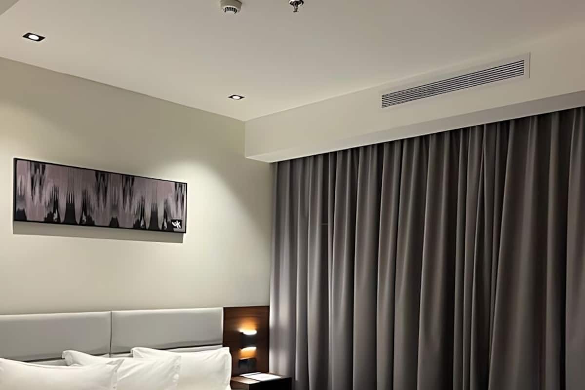 A modern hotel room with a large bed, a headboard, a bedside table with a lamp, a painting on the wall, and a window with curtains.