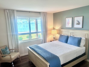A bedroom with a bed, a chair, a window with curtains, a lamp, and two wall-mounted pictures.