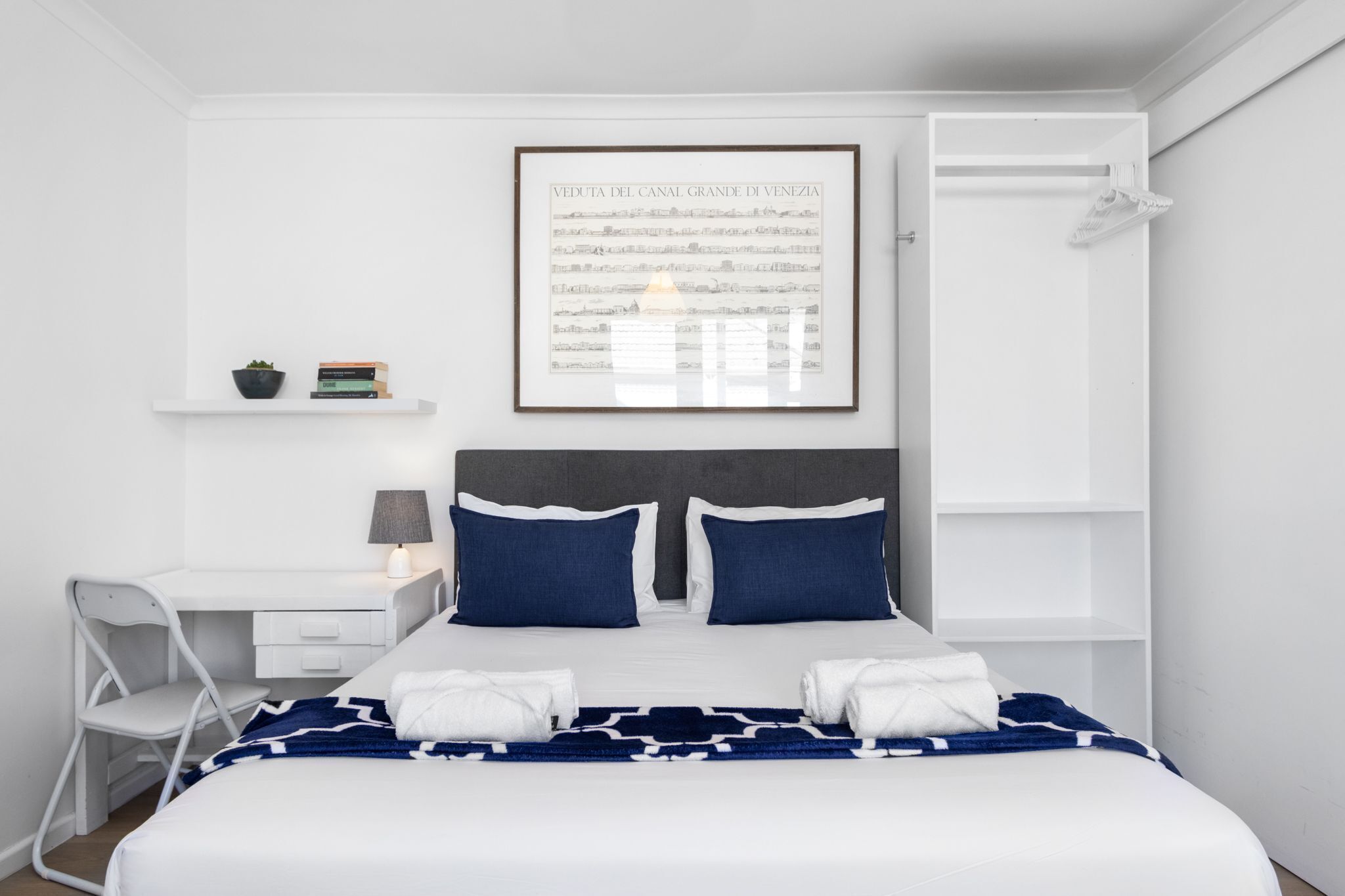 A neatly arranged hotel room with a bed, a desk, a chair, a shelf with books and a plant, and a framed picture on the wall.