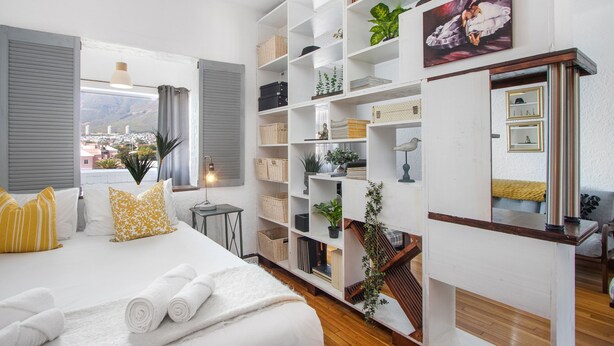 A hotel room with a bed, a large window, a bookshelf, and a view of the city. - Langholm 45 on Kloof (Cape Town)