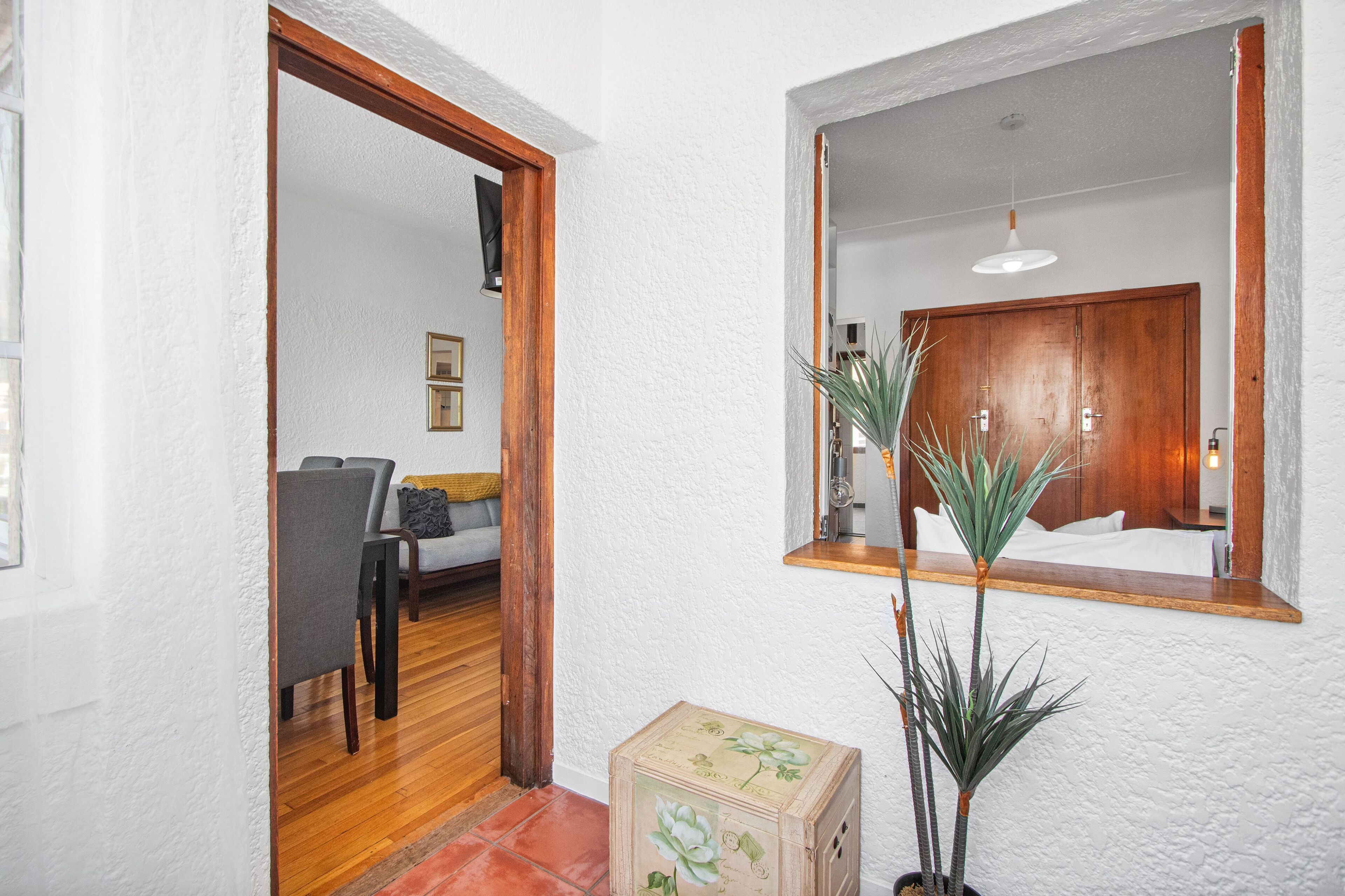 Un pasillo con piso de madera, pared blanca con textura y una puerta de madera que da a una habitación con cama, mesita de noche y espejo.