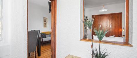 Un pasillo con piso de madera, pared blanca con textura y una puerta de madera que da a una habitación con cama, mesita de noche y espejo.