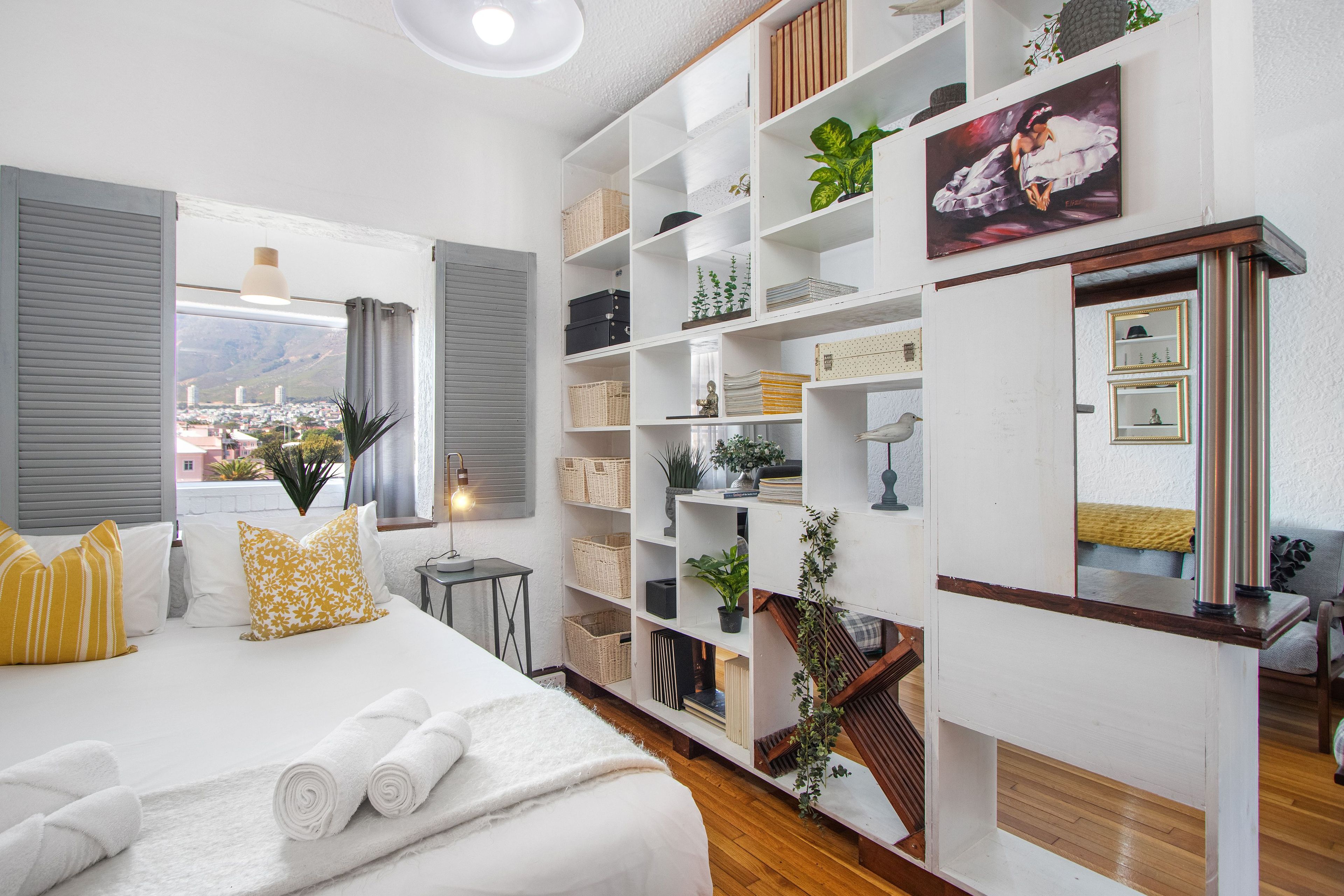A hotel room with a bed, a large window, a bookshelf, and a view of the city.