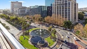 A cityscape with modern buildings, a roundabout with a fountain, and palm trees. - Fountain Suites 1102 (Cape Town)