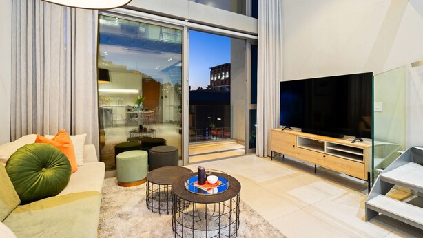 Uma sala de estar moderna com sofá, pufes, mesa de centro, televisão e porta de vidro que dá para uma varanda.