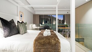 Quarto de hotel moderno com cama grande, divisória de vidro e vista da cidade ao entardecer.