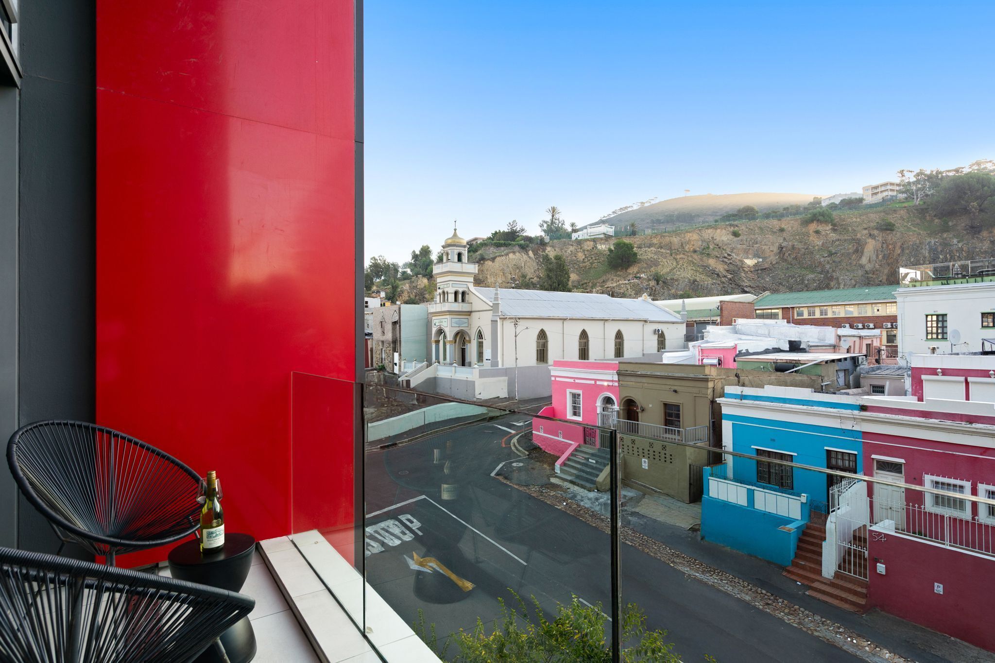 A balcony with a red wall, a black chair, and a bottle of wine.