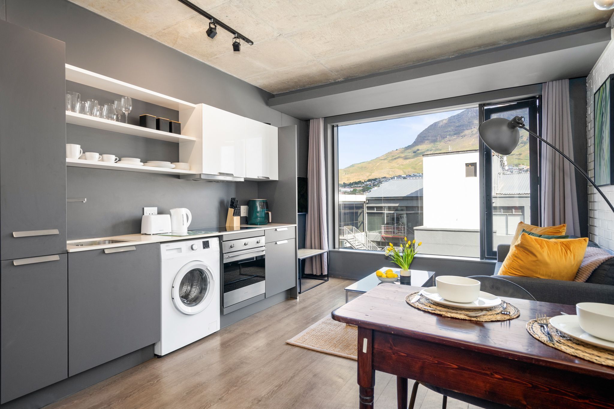 A modern kitchen with built-in appliances, a dining area with a table set for two, and a large window offering a view of a mountain and buildings.