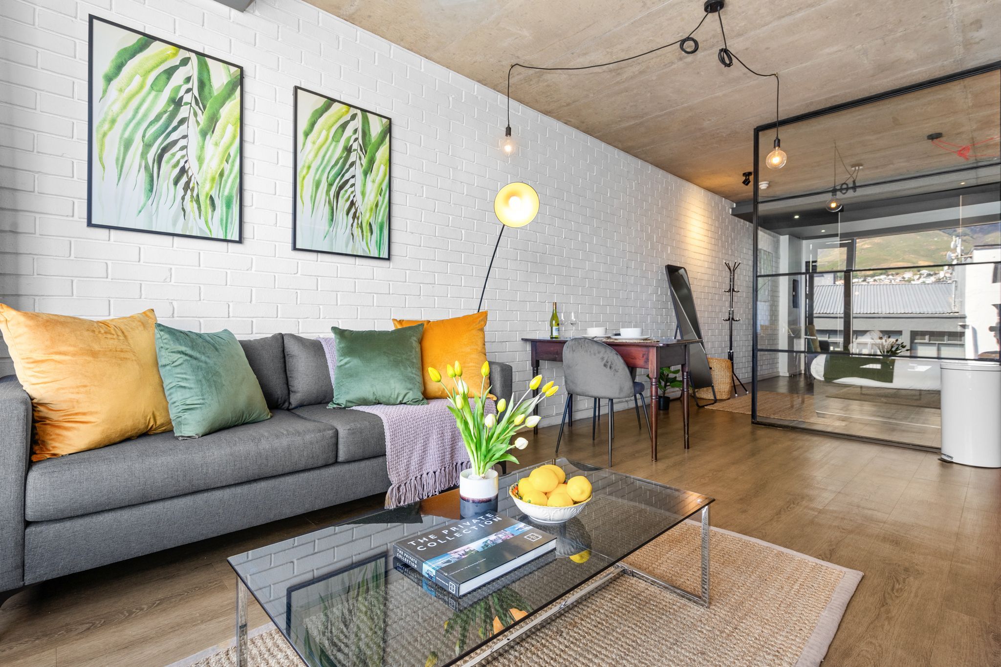 A modern living room with a grey sofa, colorful cushions, a glass coffee table with a book and a bowl of lemons, and a dining area with a glass partition.