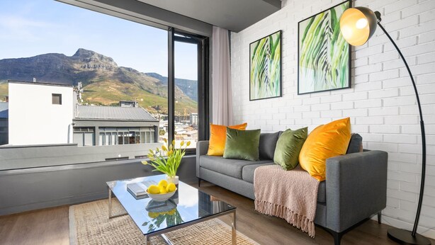 Una sala de estar moderna con un sofá gris, cojines coloridos, una mesa de centro de vidrio y vistas a montañas a través de ventanas amplias.