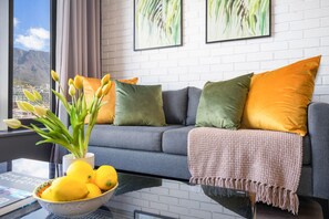 A modern living room with a grey sofa, green and orange cushions, a vase with yellow flowers, and a bowl of lemons on a glass table.