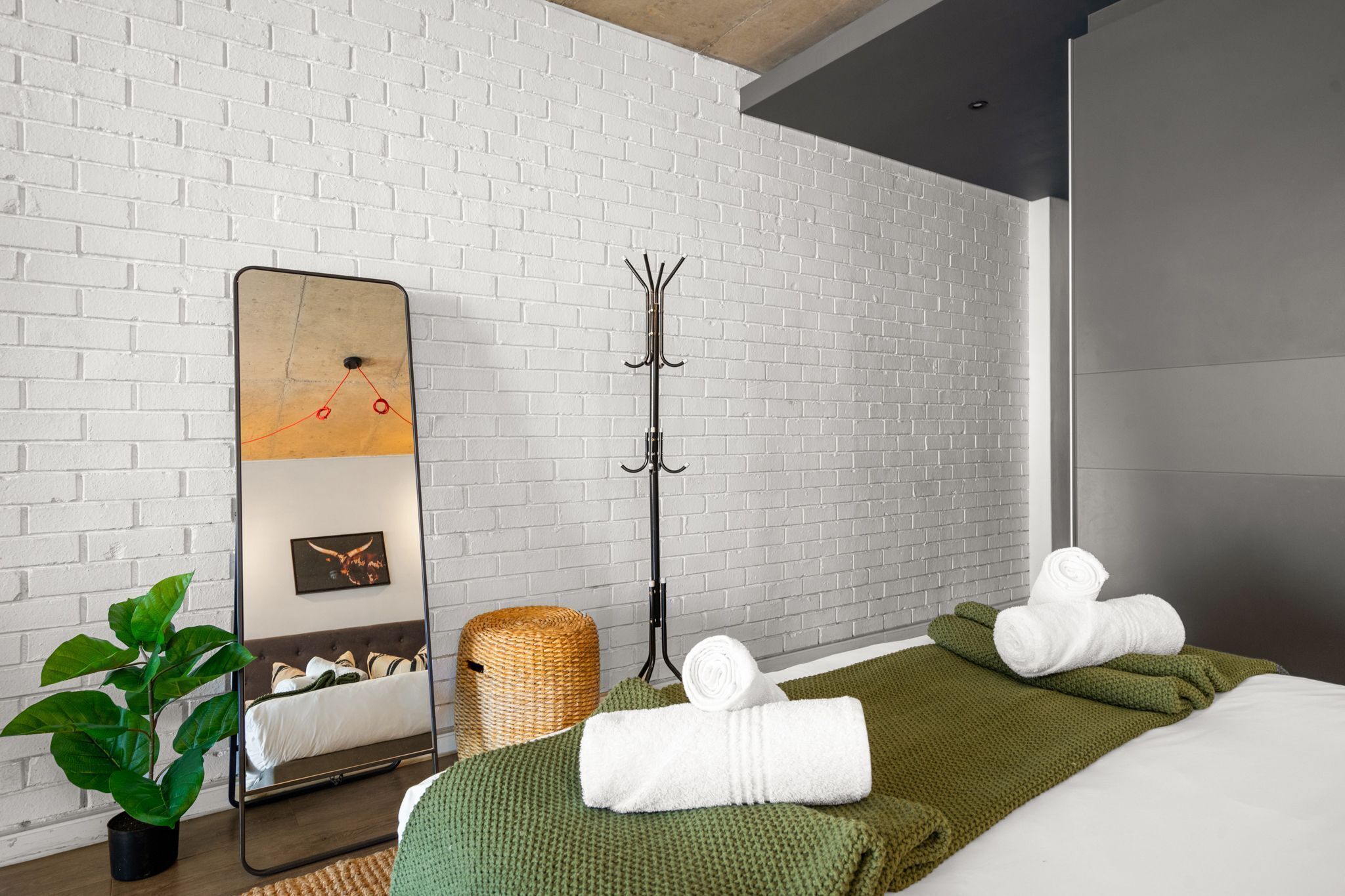 Una habitación de hotel moderna con pared de ladrillo, un espejo grande, un perchero, una cama con ropa de cama verde y blanca y una planta en maceta.