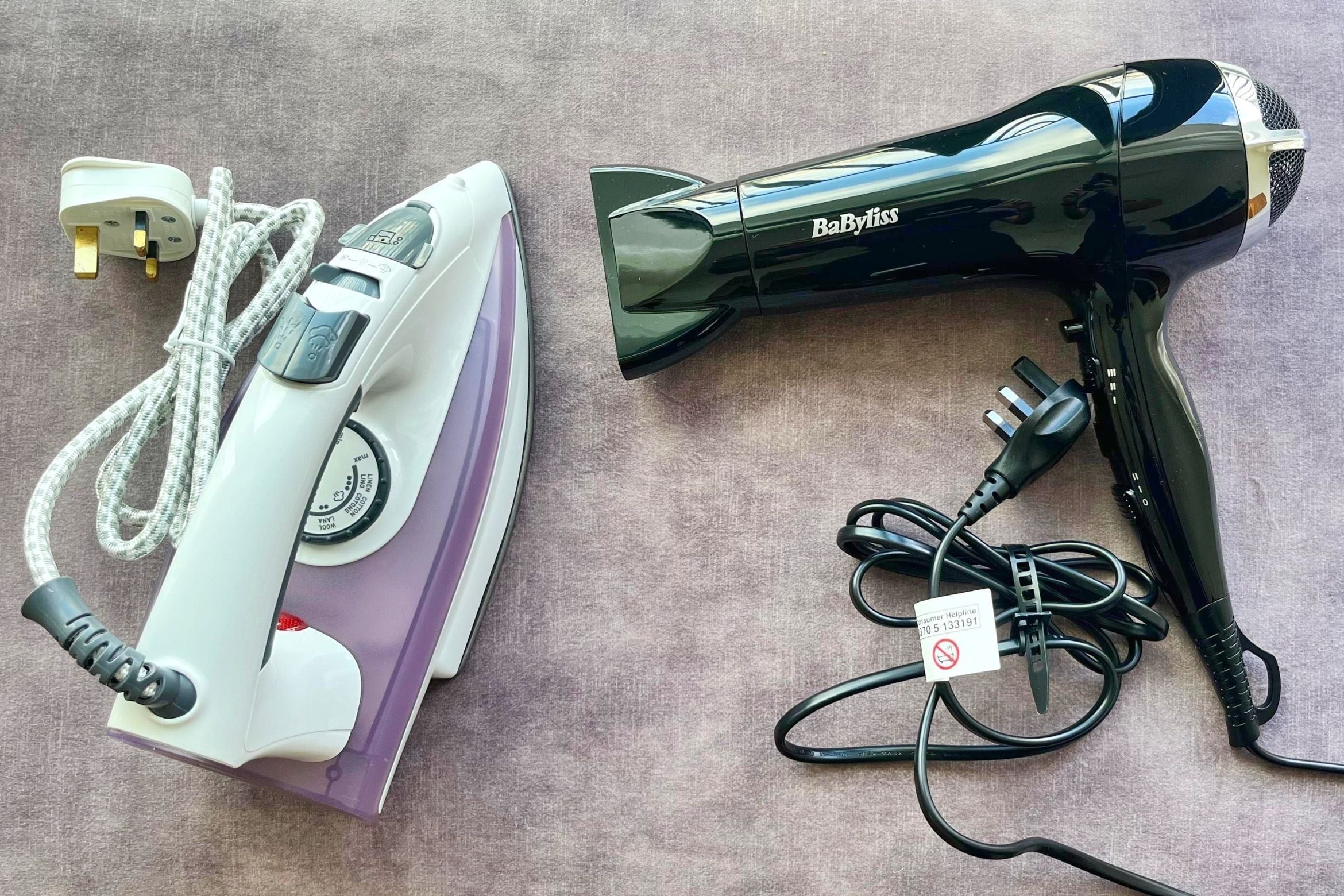 A white and black iron and a black hair dryer with a power cord and a plug on a textured surface.