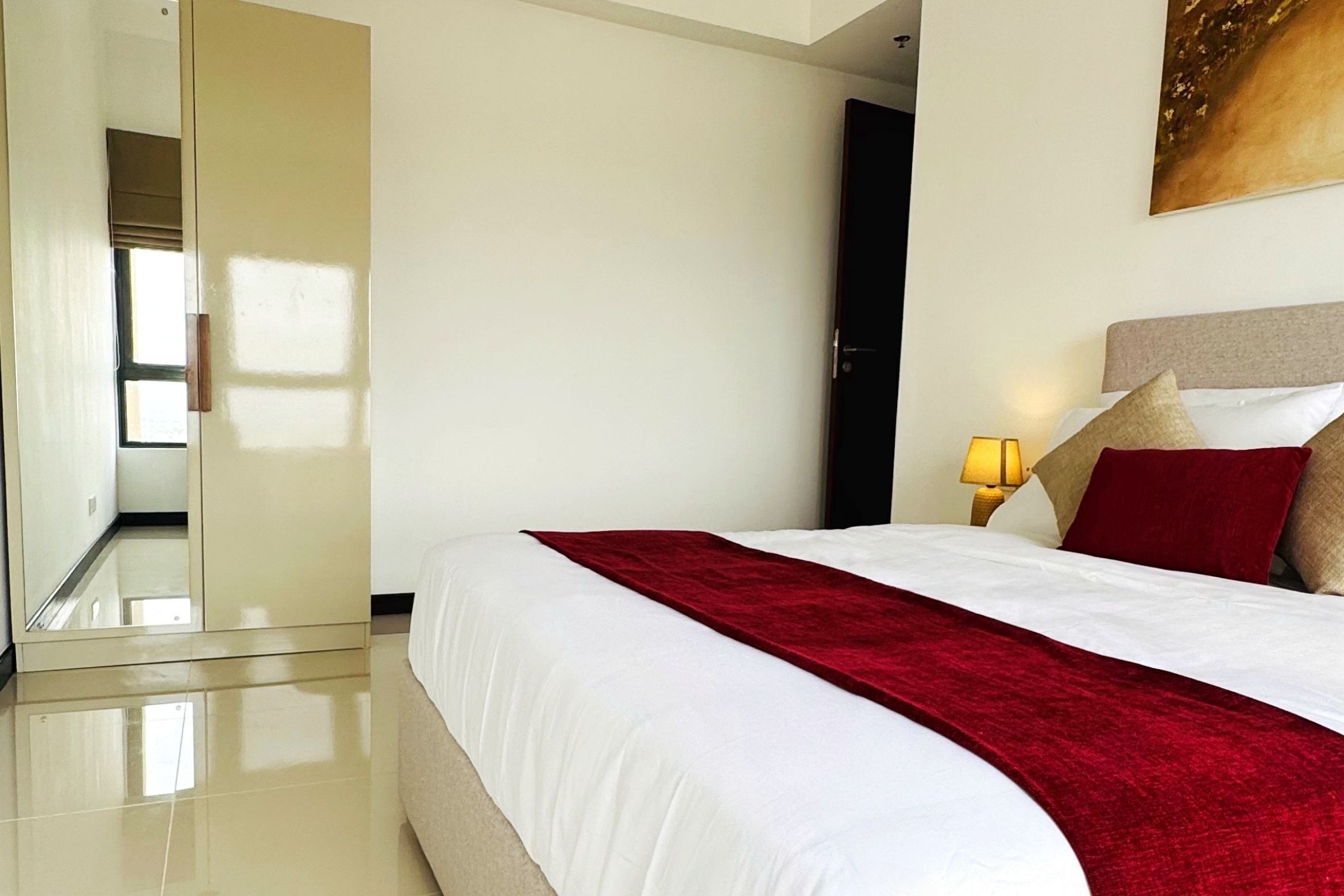 A hotel room with a large bed, a red throw blanket, a nightstand with a lamp, and a painting on the wall.