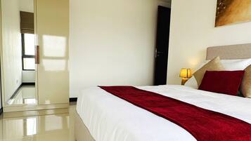 A hotel room with a large bed, a red throw blanket, a nightstand with a lamp, and a painting on the wall.