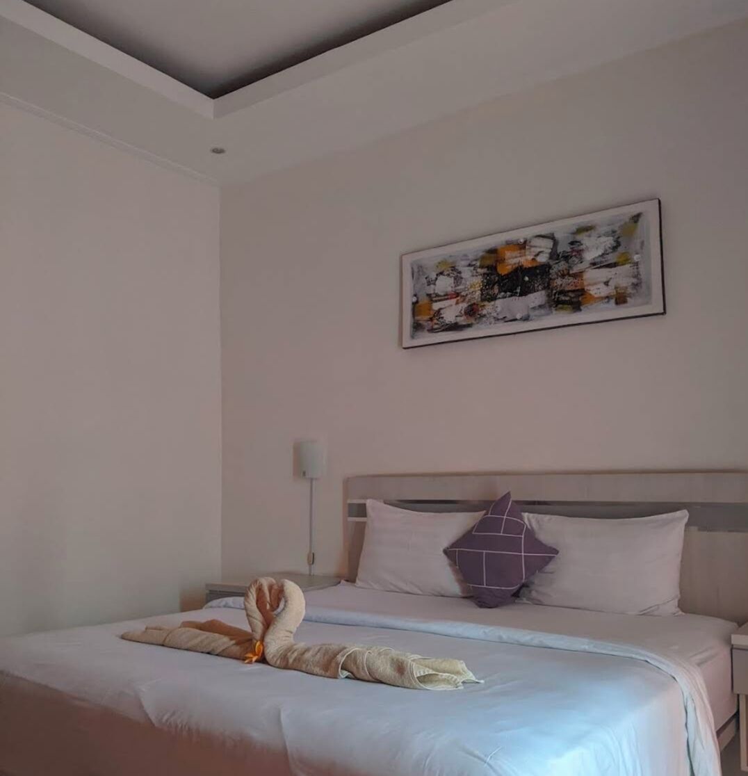 A neatly made bed with a towel and a decorative pillow, a wall-mounted abstract painting, and a bedside lamp.