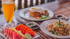 A table with a glass of beer, a plate of sliced watermelon, a plate of fried rice with a fried egg, and a plate of stir-fried noodles. - MAILAKU HOUSE (Kerobokan Kaja)