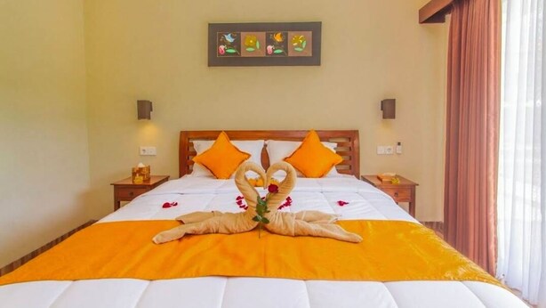 A neatly made bed with a heart-shaped towel arrangement and orange bedding, a wooden headboard, two wall-mounted lamps, and a framed artwork above the bed. - MAILAKU HOUSE (Kerobokan Kaja)
