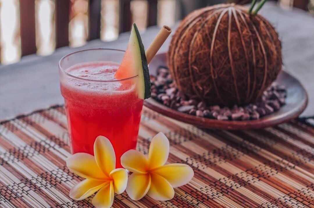 A glass of red drink with a slice of lime and a straw, a coconut, and two plumeria flowers on a woven mat.